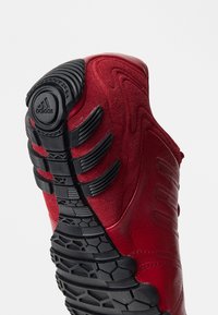 Rote Sneaker mit einer strukturierten Oberseite und schwarzen Gummielementen, einer auffälligen Sohle mit hexagonalen Mustern und einer markanten Markenkennzeichnung an der Ferse.