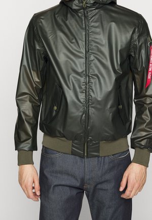 Veste à capuche vert foncé avec fermeture éclair à l'avant, poches latérales boutonnées, poignets et ourlet côtelés, portée sur un jean bleu, étiquette rouge sur la manche gauche.
