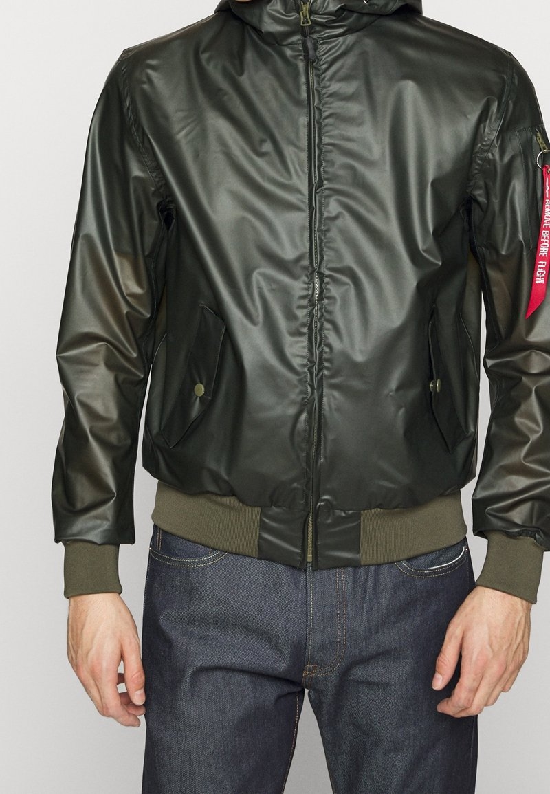Veste à capuche vert foncé avec fermeture éclair à l'avant, poches latérales boutonnées, poignets et ourlet côtelés, portée sur un jean bleu, étiquette rouge sur la manche gauche.