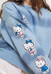 Modrá mikina s batikovaným vzorem obsahující tři barevné nášivky Hello Kitty, z nichž každá má jiný outfit a mašle, na levém rukávu.