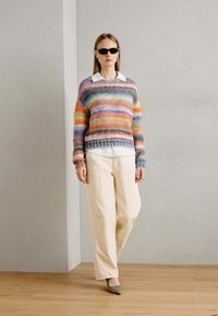 Mehrfarbiger Strickpullover mit horizontalen Streifen, darunter ein cremefarbener Kragenpullover, lockere beigefarbene Hose und dunkel gefärbte Sonnenbrille.