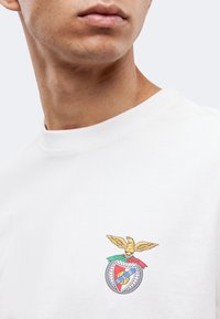 Άνδρας με λευκό πουκάμισο με έμβλημα αετού και λογότυπο ασπίδας στο αριστερό στήθος, εμφανίζεται από το λαιμό μέχρι τον άνω κορμό σε απλό φόντο.