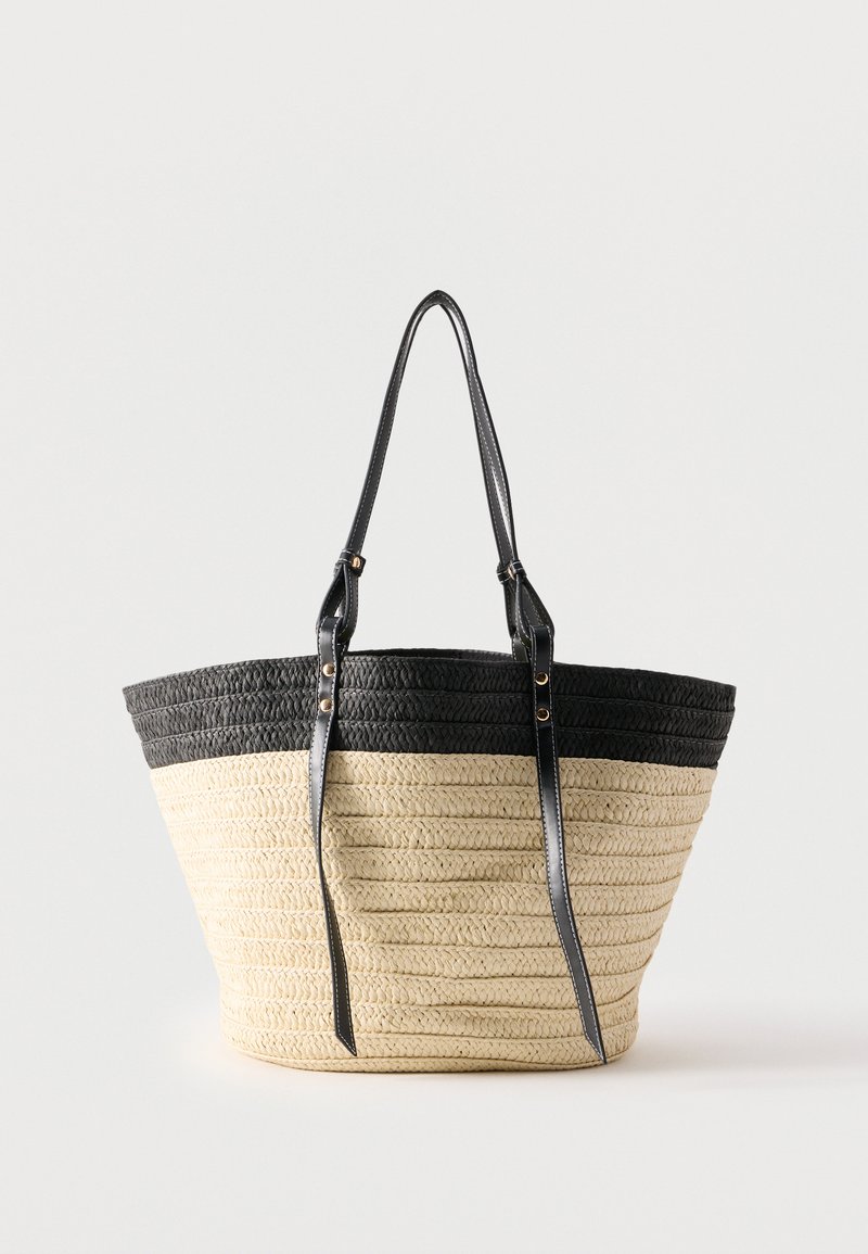 Sac fourre-tout en paille tissée avec une partie supérieure noire et de longues anses en cuir noir, présenté sur un fond clair uni.