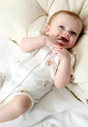 Naeratav beebi kreemika patja peal, seljas valge ühes tükis romb mustriga värviliste kuumaõhupallidega, sõrmed suu lähedal.