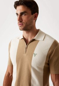 Cămășuță polo în dungi bej și crem, cu închidere prin fermoar, dintr-un material tricotat texturat, și un mic badge cu logo verde pe piept.
