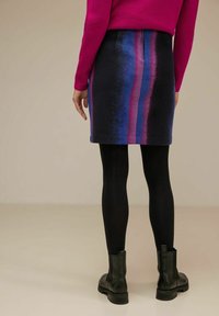 Jupe en tricot, longueur genou, avec des rayures verticales bleues et roses sur un fond sombre, assortie à des collants noirs et des bottines.