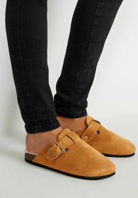 Zuecos de ante marrón con un hebilla ajustable y parte trasera abierta, combinados con jeans ajustados negros. Superficie texturizada, diseño de puntera redonda.