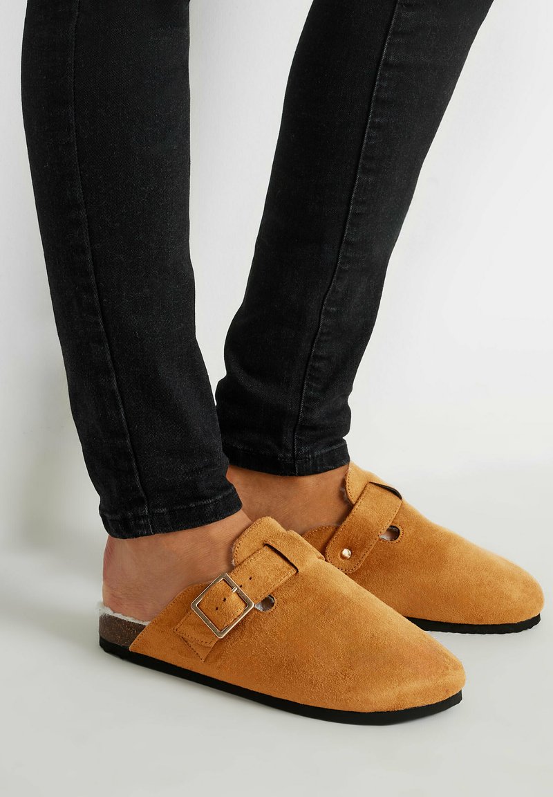 Zuecos de ante marrón con un hebilla ajustable y parte trasera abierta, combinados con jeans ajustados negros. Superficie texturizada, diseño de puntera redonda.