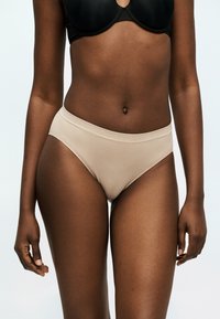 Γυναίκα που φοράει μπεζ εσώρουχο υψηλής κοπής και μαύρο σουτιέν, όρθια μπροστά σε ανοιχτό φόντο, με έμφαση στη μέση και το επάνω μέρος των μηρών.