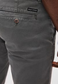 Pantalon en coton gris avec une poche arrière dotée d'un bouton marron et d'une étiquette logo noire portant l'inscription "Marc O'Polo". Le tissu présente une finition texturée.