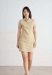 Robe polo beige à manches courtes, avec un col et un plastron à trois boutons. Matériau en coton doux, silhouette ajustée et détail de marque minimal.