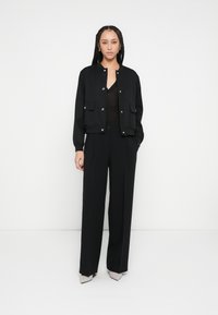 Jachetă bomber neagră cu buzunare frontale și accesorii aurii, asortată cu un top negru și pantaloni largi. Pantofii sunt ascuțiți și bej.