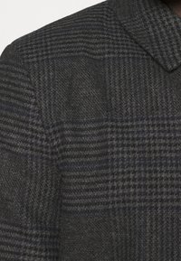 Ternede uldblazer med mørkegråt og blåt mønster, skårede revers og tekstureret finish. Nærbillede fremhæver stofdetaljer.