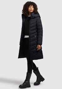Manteau matelassé noir avec capuche, présentant une fermeture éclair à l'avant, des poches latérales et une conception longueur genou. Porté avec un pantalon noir et des bottes de combat.