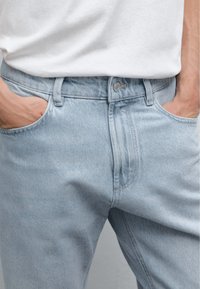 Light blue denim jeans feature a classic five-pocket design, visible stitching, and a metal button closure at the waist.