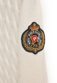Roomkleurige gebreide trui met een gestructureerd kabelpatroon. Voorzien van een rond, geborduurd Polo Club-badge in donkerblauw, goud en rode tinten.