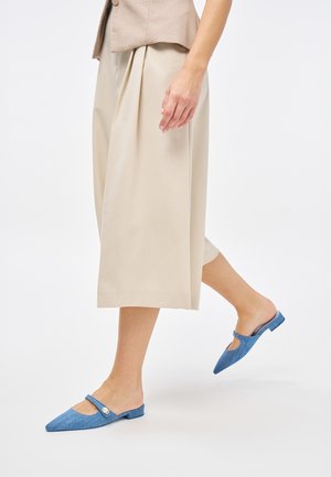 Mules en denim bleu avec un bout pointu et un détail découpé, dotées d'un petit talon et d'une finition texturée. Portées avec un pantalon beige plissé.