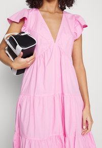 Różowa bawełniana sukienka z głębokim dekoltem w kształcie V, krótkimi falbankowymi rękawami i warstwową spódnicą. Połączona z kompaktową torebką w czarno-białym kolorze.