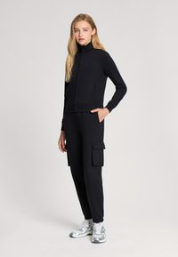 Czarna kurtka na zamek z wysokim kołnierzem, w zestawieniu z czarnymi spodniami cargo z bocznymi kieszeniami. Model nosi metaliczne trampki.