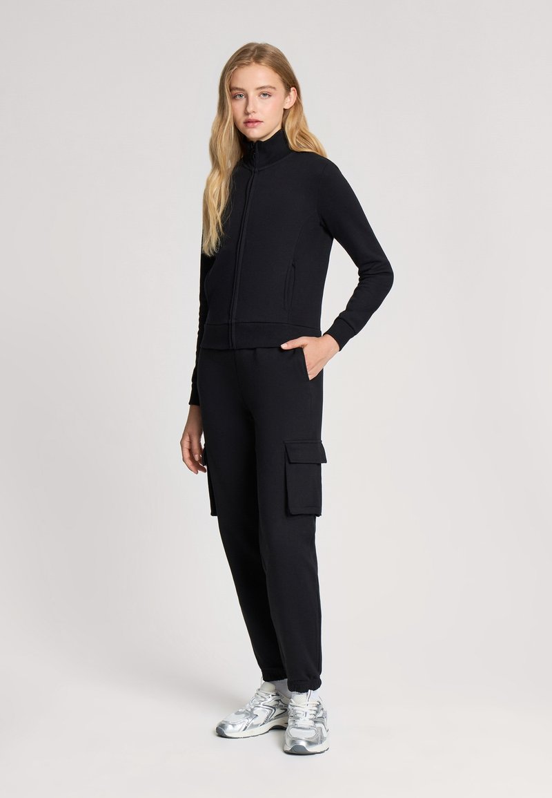 Czarna kurtka na zamek z wysokim kołnierzem, w zestawieniu z czarnymi spodniami cargo z bocznymi kieszeniami. Model nosi metaliczne trampki.