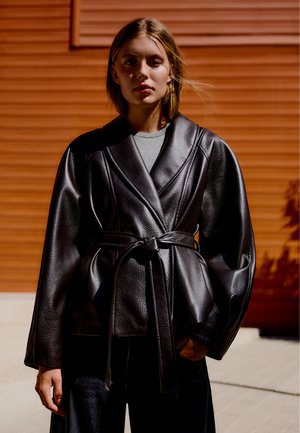 Jeune femme portant une veste en cuir noire avec ceinture et une chemise grise, debout dehors devant un mur orange au soleil.