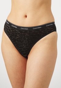 Svart blondestring med en semi-transparent tekstur, med en bred svart livline med "Calvin Klein" logo. Glatte kanter og minimal dekning.