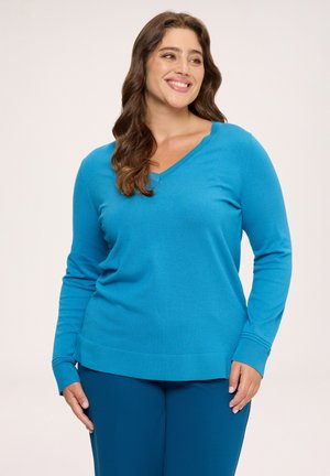 Smilende kvinde med langt brunt hår iført en lys blå V-hals sweater og matchende blå bukser, stående foran en ensfarvet baggrund.