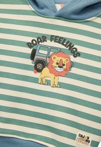 Randig hoodie i teal och krämfärg. Inkluderar en grafik av ett leende lejon med en röd man och en blå jeep, texten "ROAR FEELINGS".
