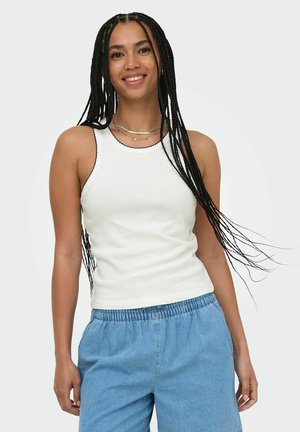 Woman with long braids smiling, wearing a white sleeveless top and loose blue pants against a plain background.