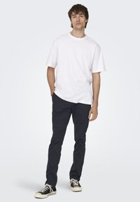 Camiseta de algodón blanca de manga corta, combinada con pantalones de algodón azul marino oscuro y zapatillas de lona negras, con un fondo gris claro.