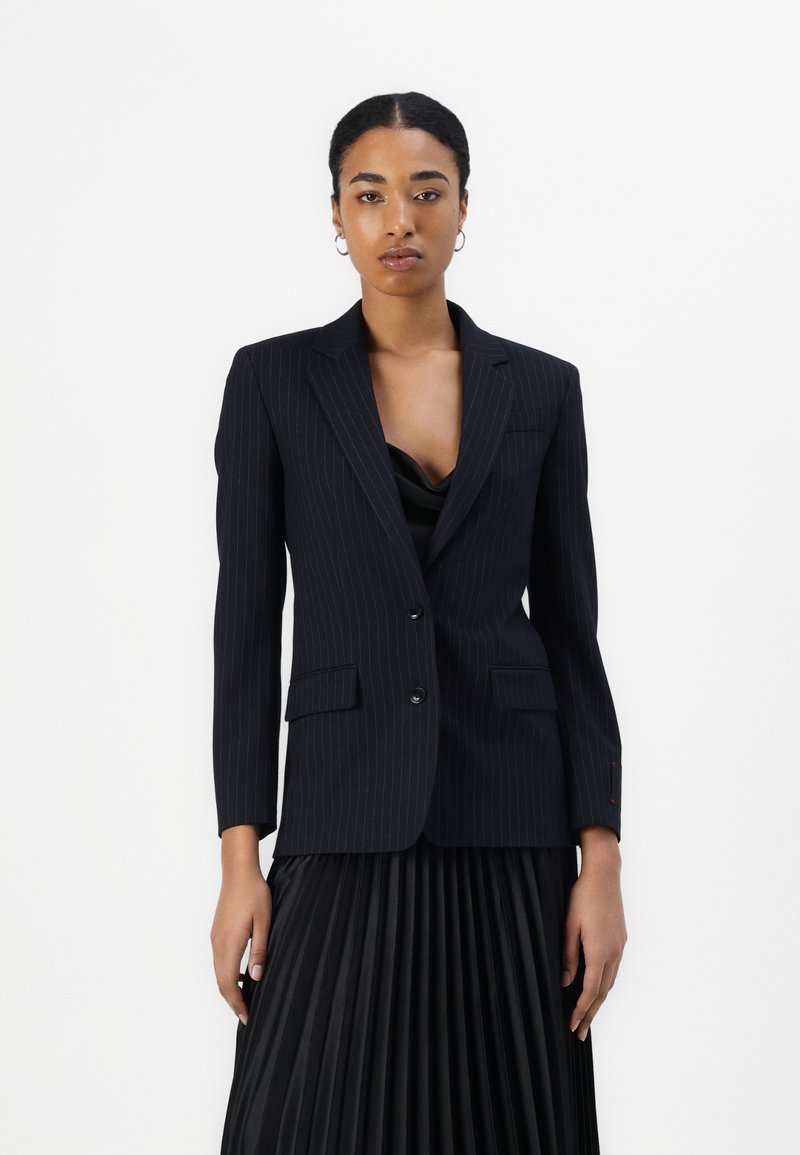 Dunkel gestreifter Blazer mit taillierter Passform, Ein-Knopf-Verschluss und zwei vorderen Taschen, kombiniert mit einem plissierten schwarzen Rock.