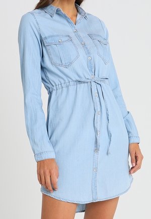 Femme portant une robe chemise en jean bleu clair à boutons, manches longues, poches poitrine et ceinture nouée à la taille.