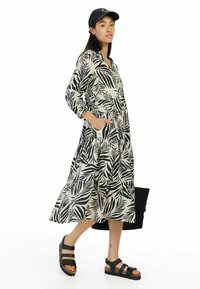 Langes, gestuftes Kleid aus leichtem Stoff mit einem schwarz-weißen tropischen Blattmuster. Begleitet von schwarzen Sandalen und einer Mütze.