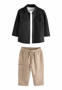 Schwarze Knopfleistenjacke mit zwei Brusttaschen, kombiniert mit beigen Jogginghosen mit elastischem Bund und Seitentasche.