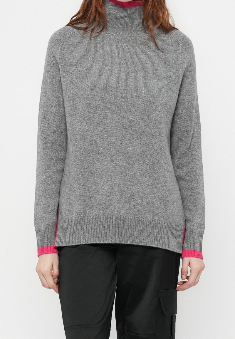 Personne portant un pull à col roulé en tricot gris avec des poignets roses et un pantalon noir avec une poche latérale, debout devant un fond blanc.