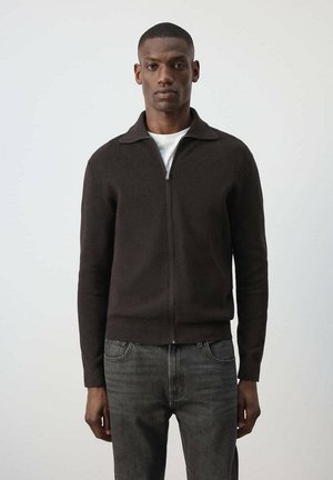 Hombre que lleva un cárdigan marrón oscuro con cremallera sobre una camisa blanca y vaqueros grises, de pie frente a un fondo claro y liso.