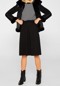 Schwarzer plissierter Rock mit hoher Taille, kombiniert mit einem gemusterten Oberteil und schwarzen Schnürstiefeletten. Der Mantel hat einen Besatz aus Kunstfell.