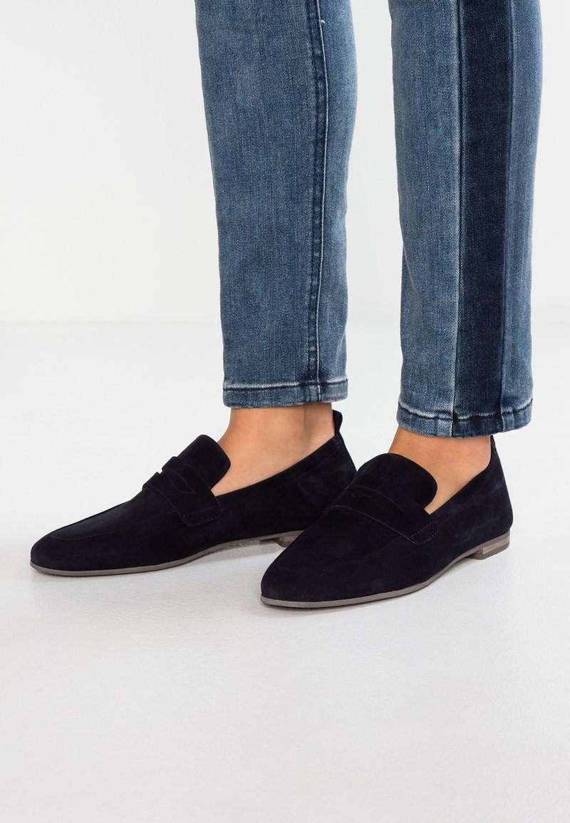Personne portant des mocassins en daim bleu foncé et un jean bleu raccourci debout sur un sol blanc.