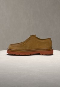 Ανδρικό καφέ σουέντ παπούτσι με κορδόνια και παχύ πορτοκαλοκαφέ ελαστικό πέλμα σε φωτεινή επιφάνεια με γκρι φόντο.