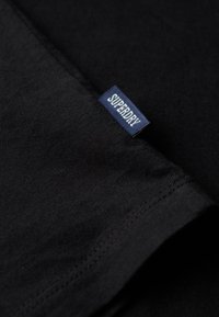 Gros plan d'une couture en tissu noir avec une petite étiquette bleue affichant le texte blanc «SUPERDRY».