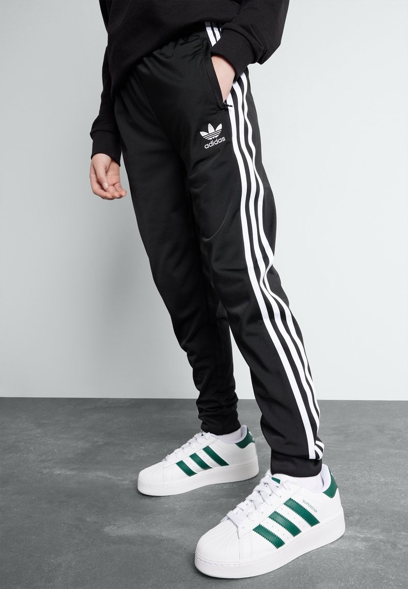 Personne portant un pantalon de jogging noir Adidas avec des rayures blanches et des baskets Adidas Superstar blanches avec des rayures vertes, se tenant sur un sol gris.