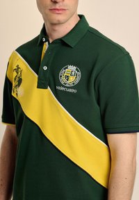 Camisa polo verde con una franja diagonal amarilla que presenta un logo bordado y un motivo de caballo. Manga corta, cuello y tapeta de dos botones.