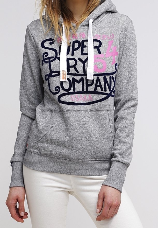 Personne portant un sweat à capuche gris avec des cordons blancs et le texte « Superdry Company » en bleu marine et rose, associé à un pantalon blanc.