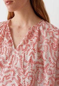 Blusa bianca con motivo floreale rosso, con scollatura a V e dettagli arricciati attorno al collo. Tessuto leggero.