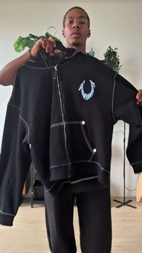 Fekete kapucnis pulóver pamut keverékből, kontrasztos fehér varrással, a mellkason kék grafikával és elülső, patentos zsebekkel.