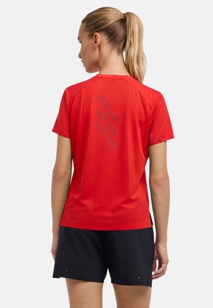 Vrouw met blonde paardenstaart van achteren te zien, draagt een rood T-shirt met korte mouwen met kleine grijze patronen en zwarte korte broek, staand tegen een effen achtergrond.
