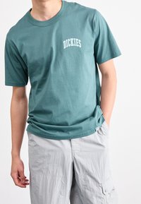 T-shirt en coton bleu sarcelle à manches courtes et col rond, avec le logo blanc "DICKIES" sur la poitrine. Texture douce et coupe décontractée.