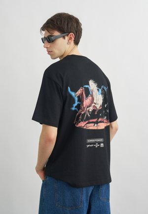 Νεαρός άνδρας που φοράει μαύρα γυαλιά ηλίου και μαύρο T-shirt με γραφικό τρεις άλογα που τρέχουν στην πλάτη, συνδυασμένο με μπλε τζιν.