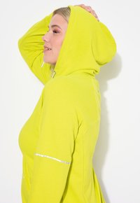 Felpa con cappuccio verde lime dal taglio morbido, con accenti riflettenti sulle maniche e una superficie morbida e liscia.