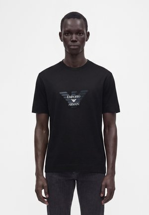 Άνδρας που φοράει μαύρο t-shirt Emporio Armani με εκτύπωση λογότυπου, όρθιος μπροστά σε απλό λευκό φόντο.